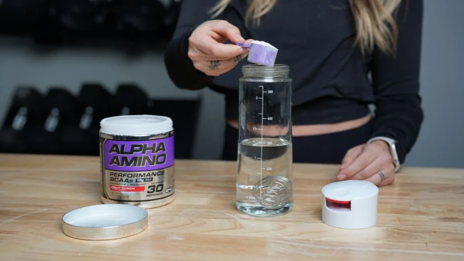 A person in a black long-sleeve top is scooping powder from a container labeled Alpha Amino Performance BCAAs into a clear water bottle with measurement markings. The bottle sits on a wooden table beside the container lid.