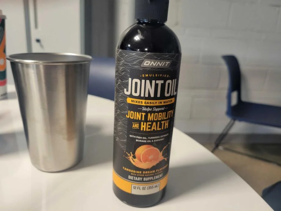 A bottle of  Onnit Joint Oil is next to a stainless steel cup on a tabletop.