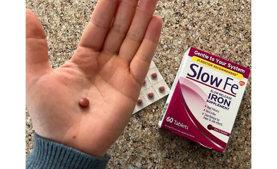 Close-up of a hand holding a single small red tablet next to an aluminum sealed pack of more tablets and a box of Slow Fe Slow Release 45mg Iron supplements.