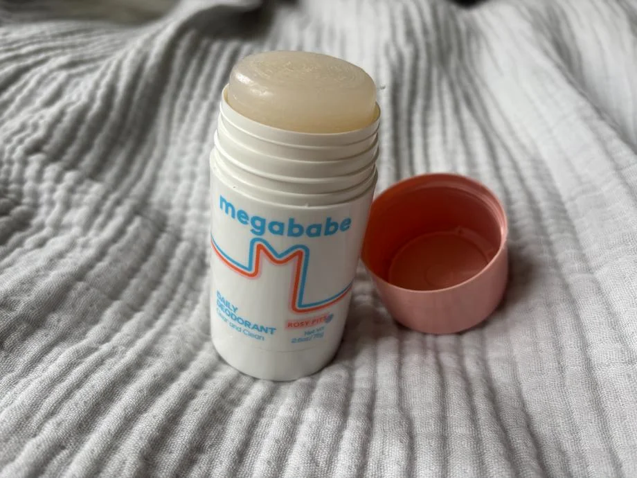 An opened stick of Megababe Rosy Pits Daily Deodorant is staged on a rumpled bedspread.