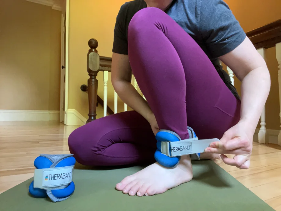A shot from the shoulders down of a woman putting on the blue Theraband ankle weights while in a kneeling position