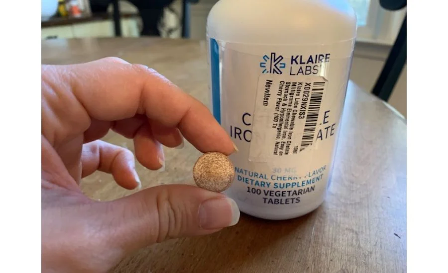 Close-up of a hand holding a large and round brown chewable tablet next to a white bottle of Klaire Labs Chewable Iron Chelate supplements.
