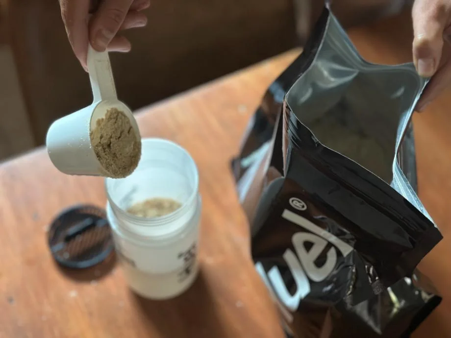 A woman pouring a scoop of Huel Complete Black protein powder into a shaker cup.