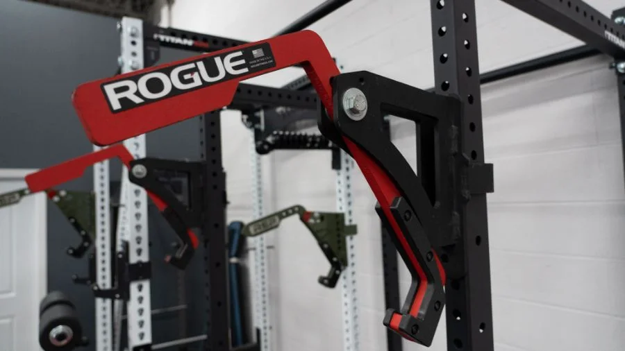 Close-up of a red and black gym equipment accessory with the text ROGUE in white. It is attached to a dark metal frame, with another similar piece in the background. The surrounding walls and equipment are mostly gray and black.
