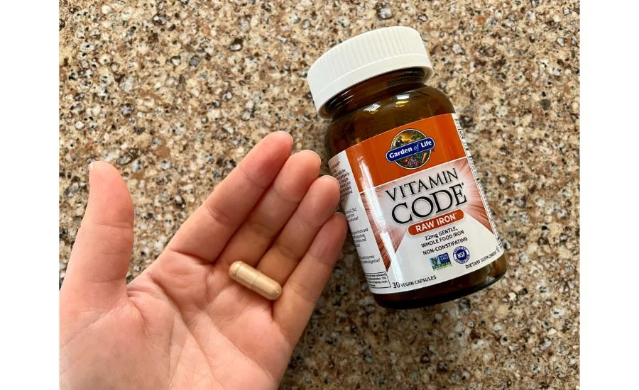 Close-up of a hand holding a single large capsule next to a brown bottle with red labeling of the Garden of Life Vitamin Code raw iron supplements.