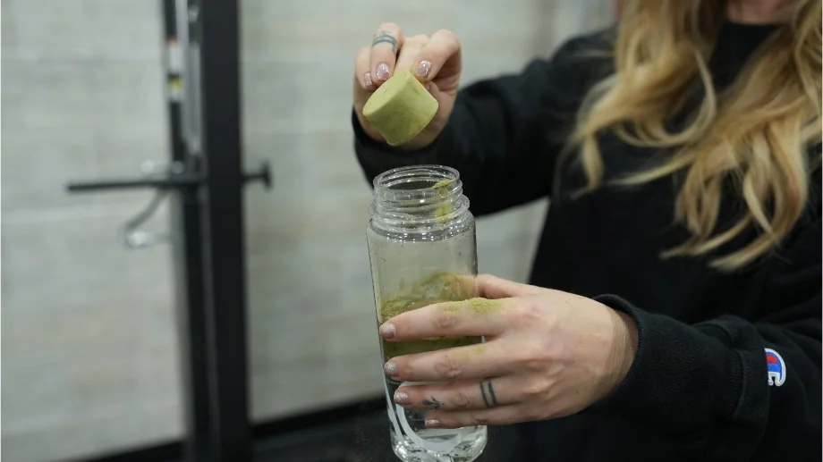 A woman pours a scoop of AG1 greens powder into a shaker bottle