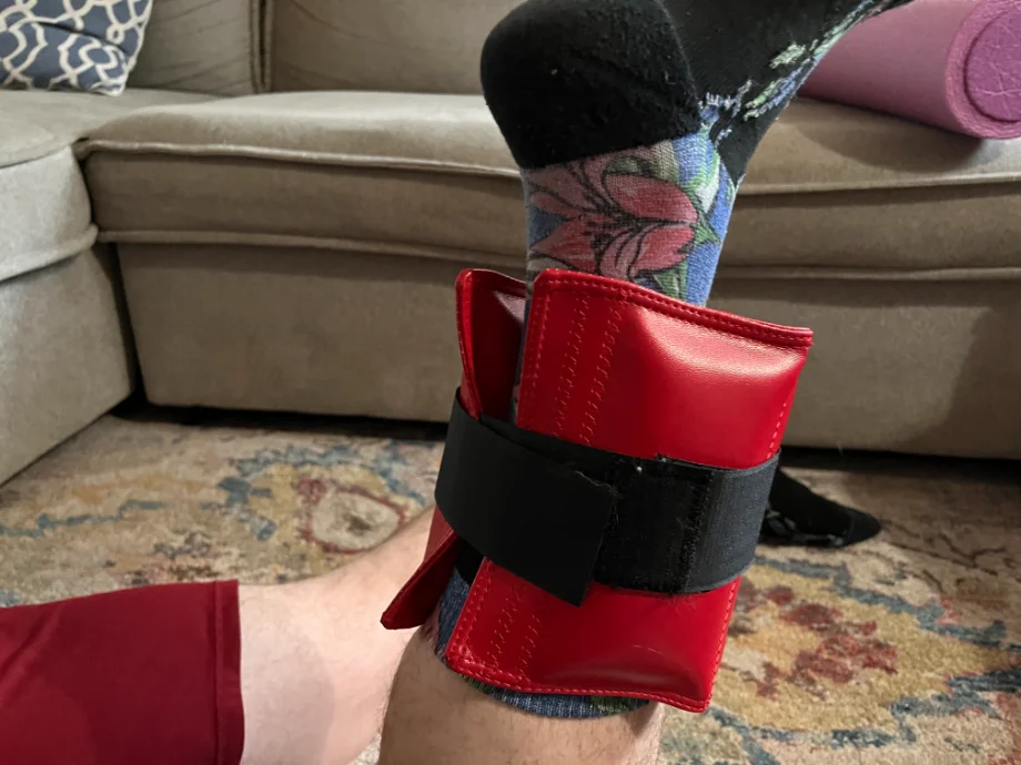 A Close-up shot of a man's left ankle while wearing The Cuff ankle weight in red, strapped on in prone position