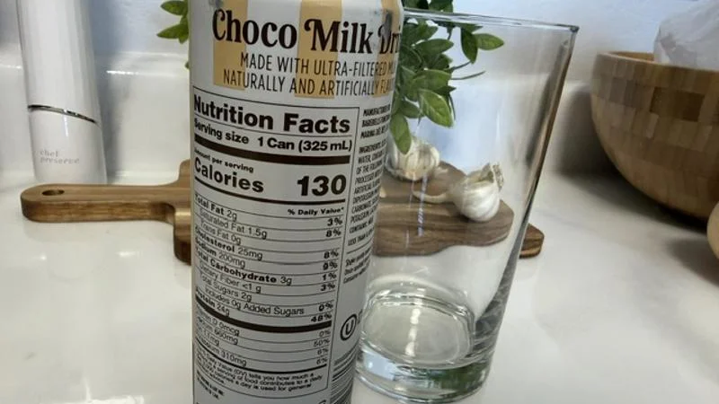 An empty glass behind an unopened can of Barebells Choco Milk Drink with the Nutrition Facts label showing.