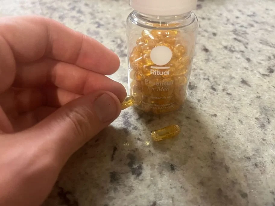 Close-up of a hand holding a single gel capsule filled with a yellowish liquid, next to a loose capsule sitting on the counter and a clear bottle of Ritual Essential Multivitamin for Men 18+.