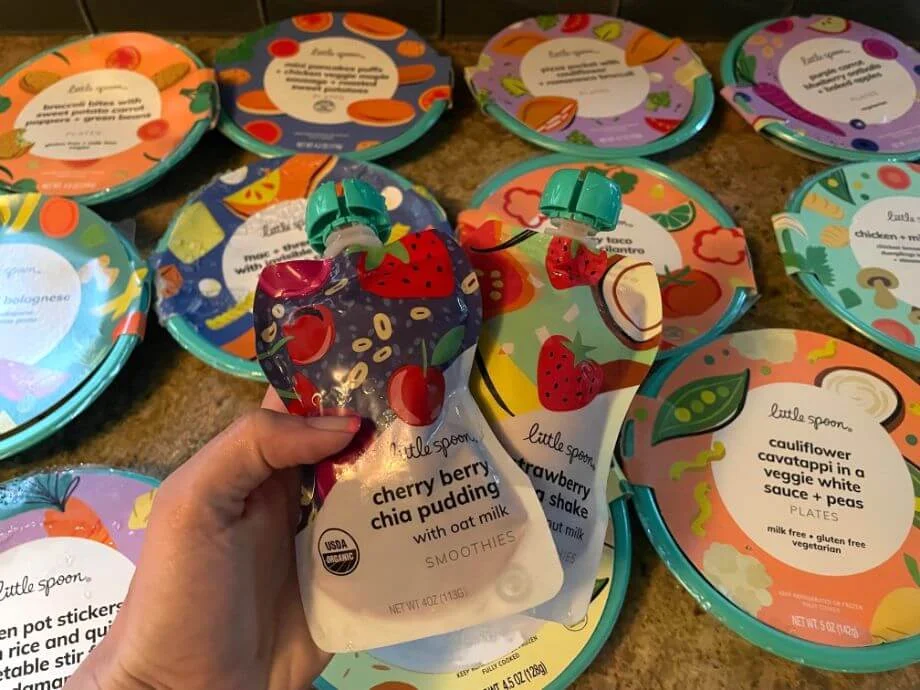 A person holds two smoothie pouches in front of an array of plates from Little Spoon