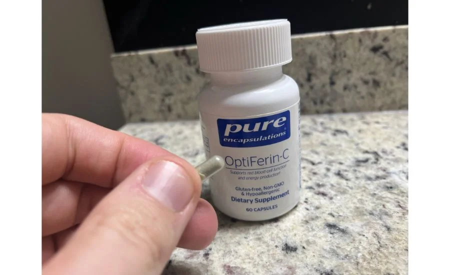 Close-up of a hand holding a single capsule filled with a whiteish powder between two fingers, next to a white bottle of Pure Encapsulations OptiFerin-C dietary supplements.