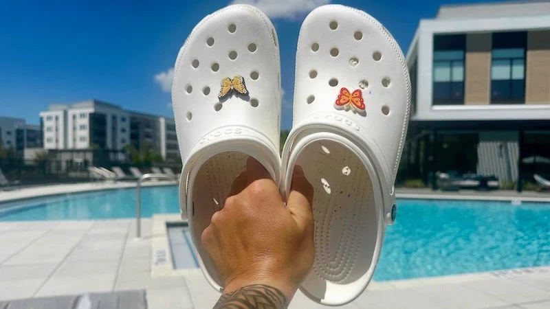 A person holding up a pair of Croc Classic Clogs with a swimming pool in the background.