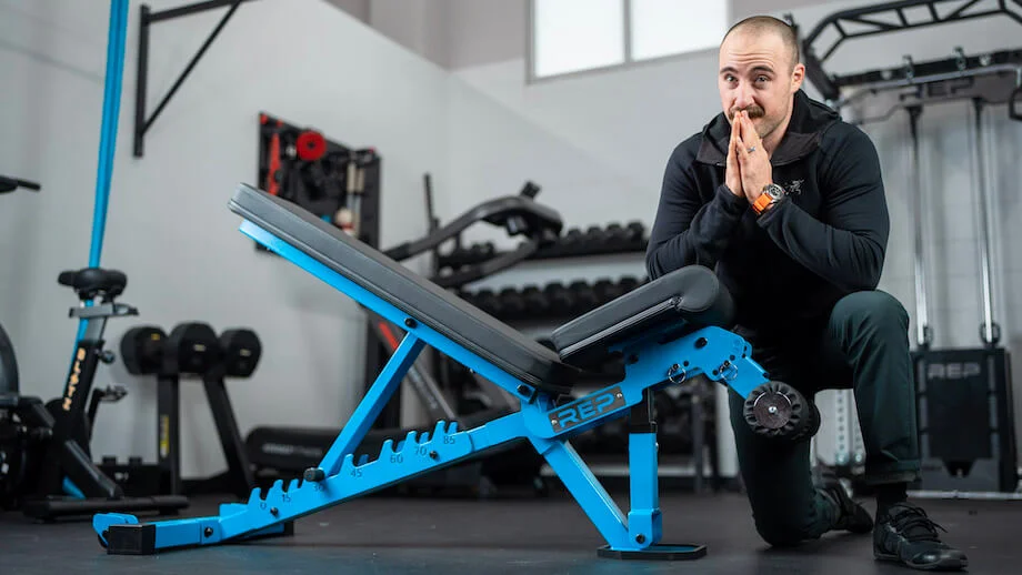 Coop kneels next to a REP Fitness AB-3000 2 weight bench. The bench is blue with a black pad.
