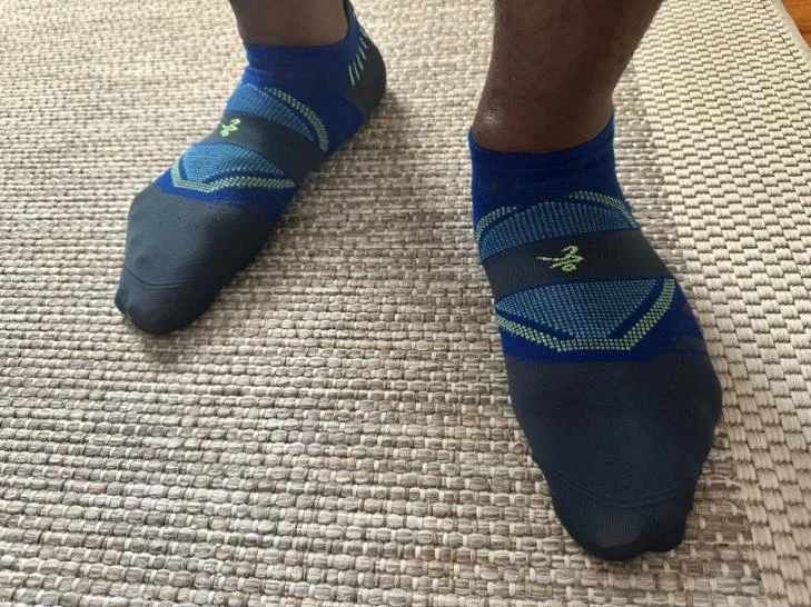 Close-up of a person’s feet in blue and black patterned Balega Hidden Dry No Show Athletic Socks on a textured carpet surface.