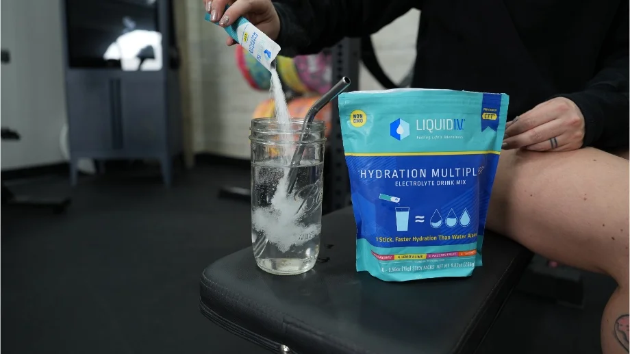 A close-up image of a woman's hand pouring a packet of Liquid IV into a water bottle that's standing next to the electrolyte container