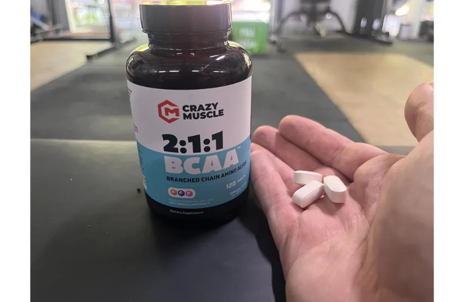 A person holds four white tablets in their hand. In the background, there is a bottle labeled Crazy Muscle 2:1:1 BCAA Branched Chain Amino Acids with 120 tablets, placed on a dark surface in a gym setting.