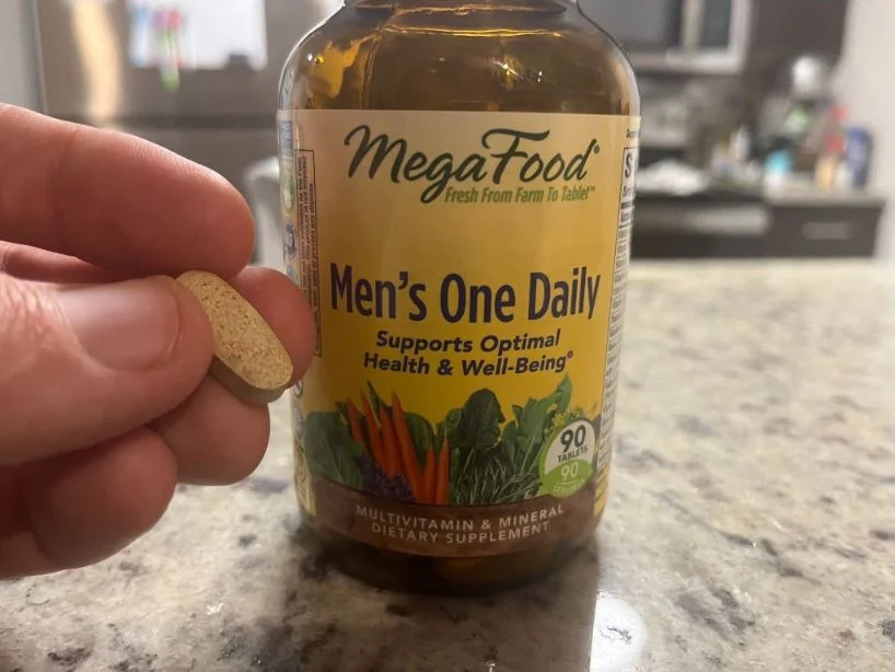 Close-up of a hand holding a large oval tablet in front of a brown colorfully labeled bottle of MegaFood Men's One Daily Multivitamin. food multivitamin capsule