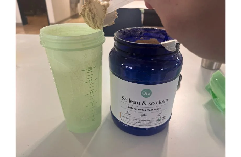 Someone dumping a scoop of Ora Organic plant protein into a light green shaker cup. The dark blue container of protein powder is next to the cup.