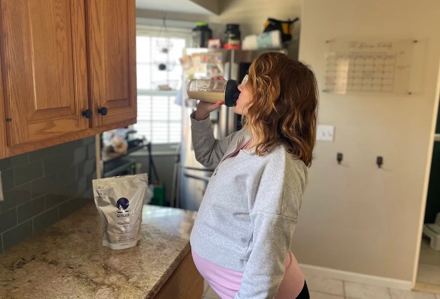 A pregnant woman standing in a kitchen drinking Ritual Essential protein powder for pregnancy out of a clear shaker bottle