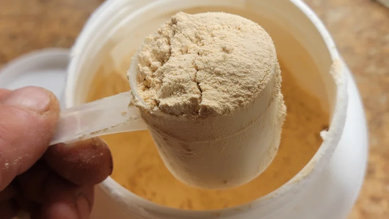 A hand lifts a scoop of TumLove Gut-Friendly Protein Powder from the opened container below