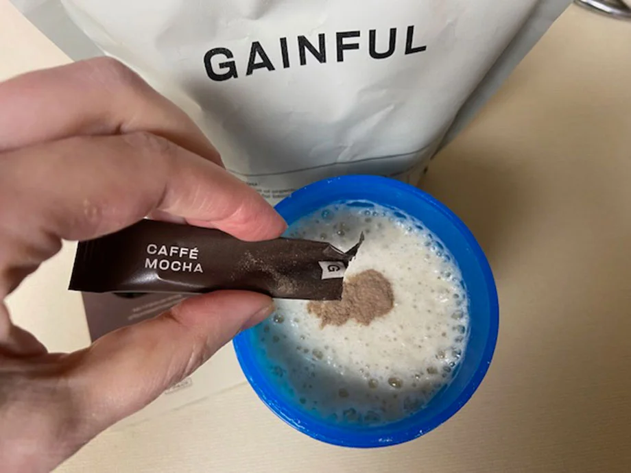 Someone pouring Caffe Mocha flavor boost into Gainful vegan protein powder in a blue shaker cup