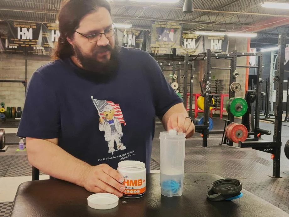 A man pours a scoop of Blonyx HMB+ Creatine into a shaker cup while inside of a gym