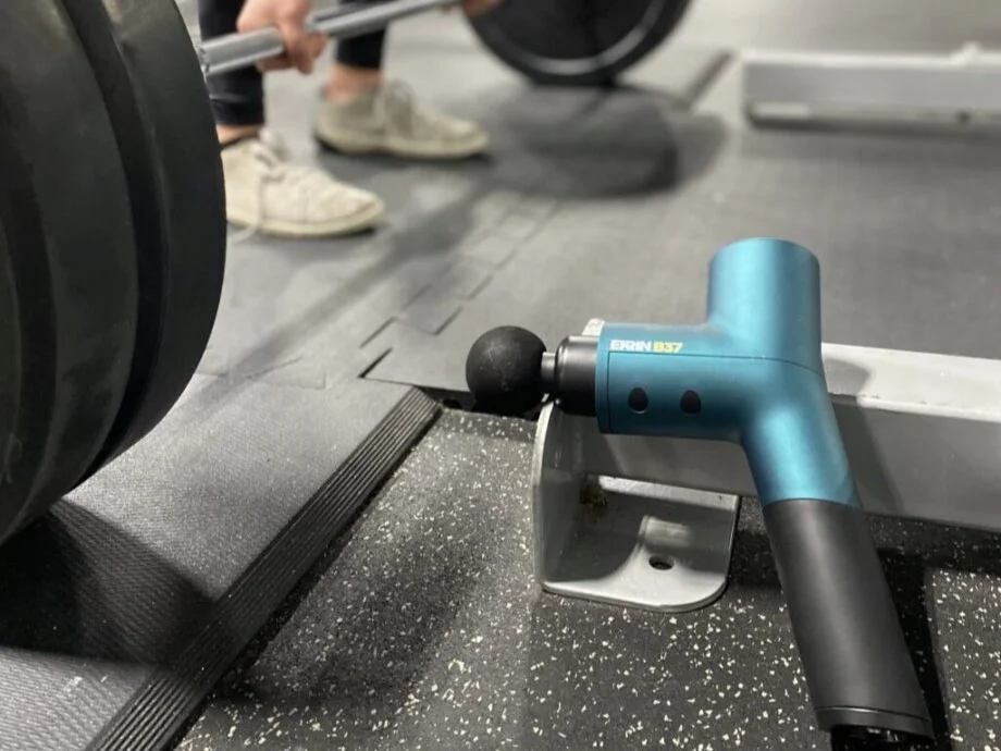 A person deadlifts with the Ekrin Athletics B37 massage gun placed in front of them, resting on the feet of a squat stand
