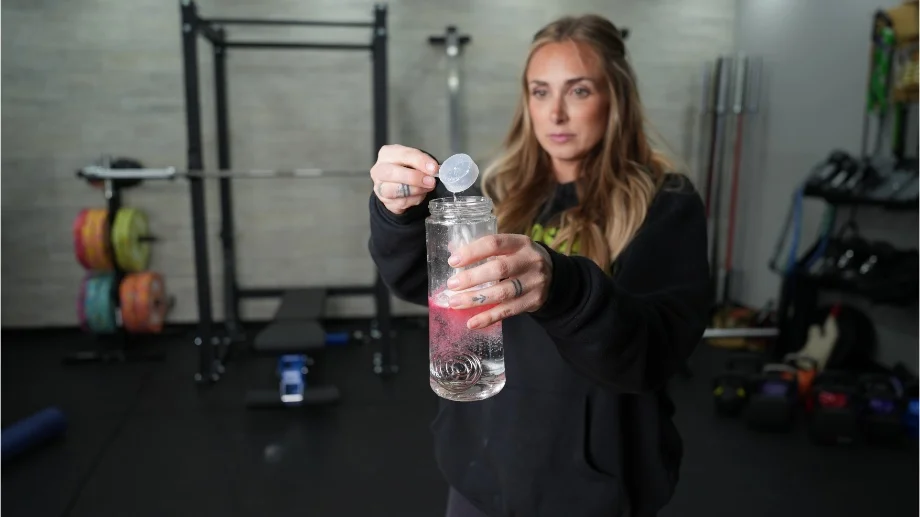 A woman in a gym is pouring Optimum Nutrition Gold Standard Pre-Workout into a clear shaker bottle. The water is turning red from the powder.