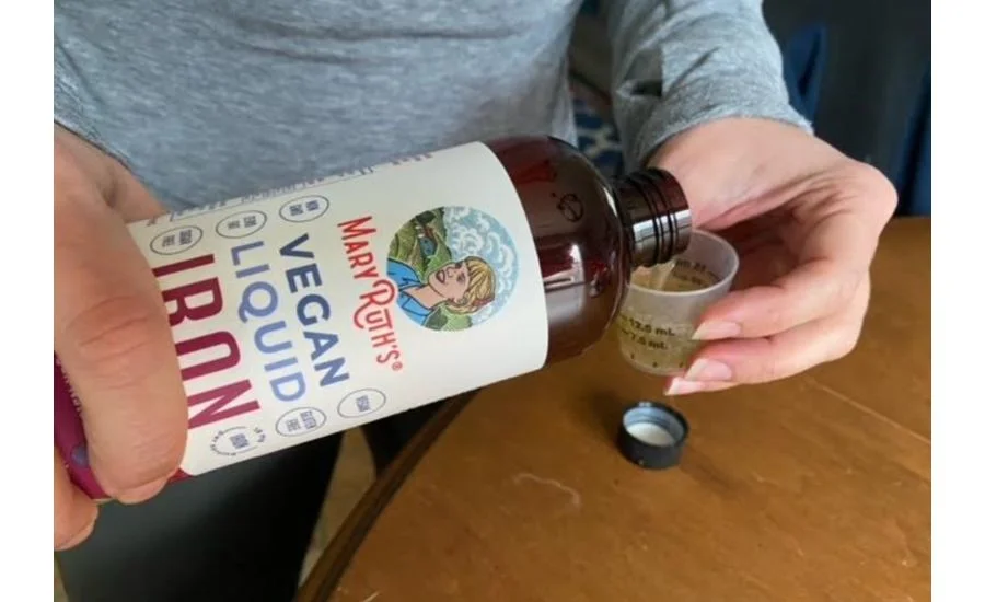 Close-up of a person pouring a yellowish liquid out of a brown bottle of Mary Ruth's Organics Liquid Iron supplement with vintage branded imagery into a small measuring cup.