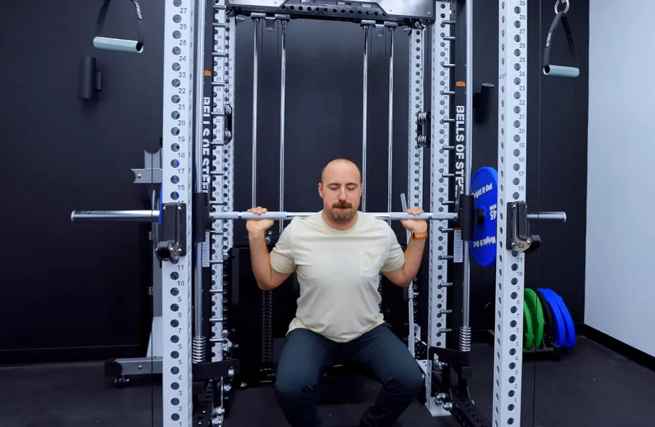 A man is shown squatting in the Bells of Steel Smith Machine Attachment