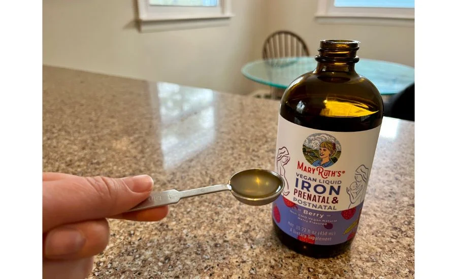 A hand holding a metal measuring spoon filled with a yellowish liquid, next to a brown glass bottle of Mary Ruth Organics Liquid Prenatal & Postnatal Iron.