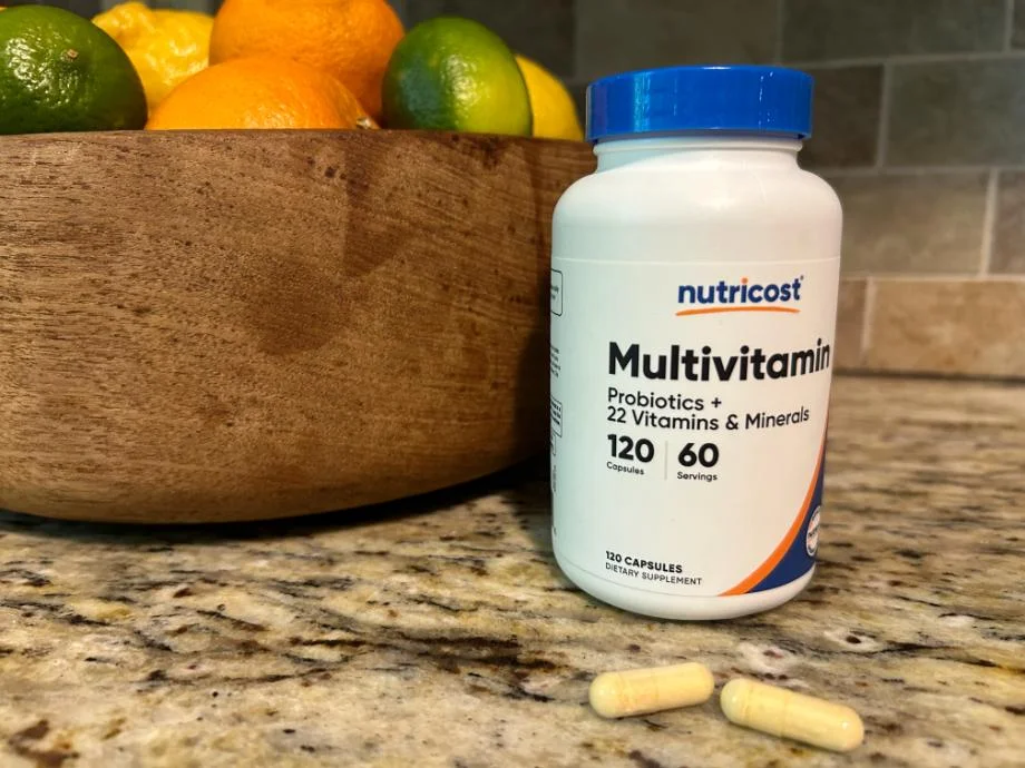 Close-up of two yellowish capsules sitting on a counter in front of a bottle of Nutricost Multivitamins with a blue lid.