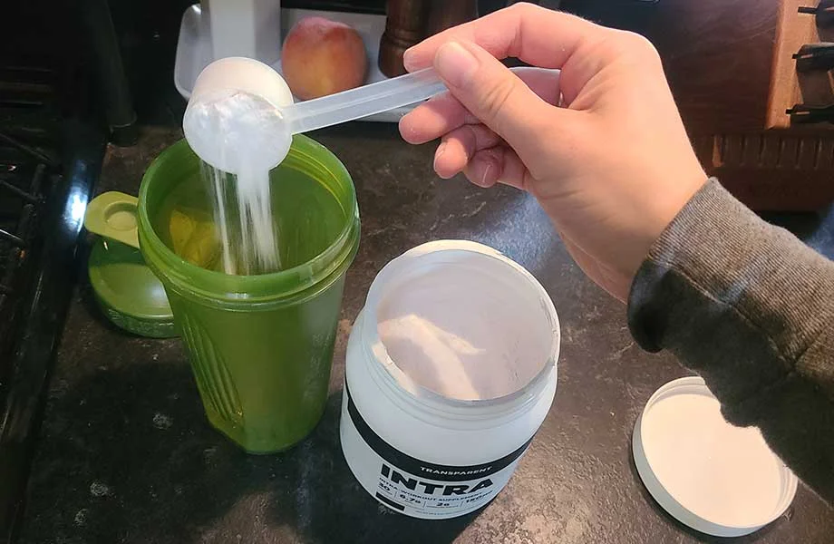 A hand is scooping white powder from a container labeled INTRA into a green shaker cup on a kitchen counter. A peach and other kitchen items are visible in the background.