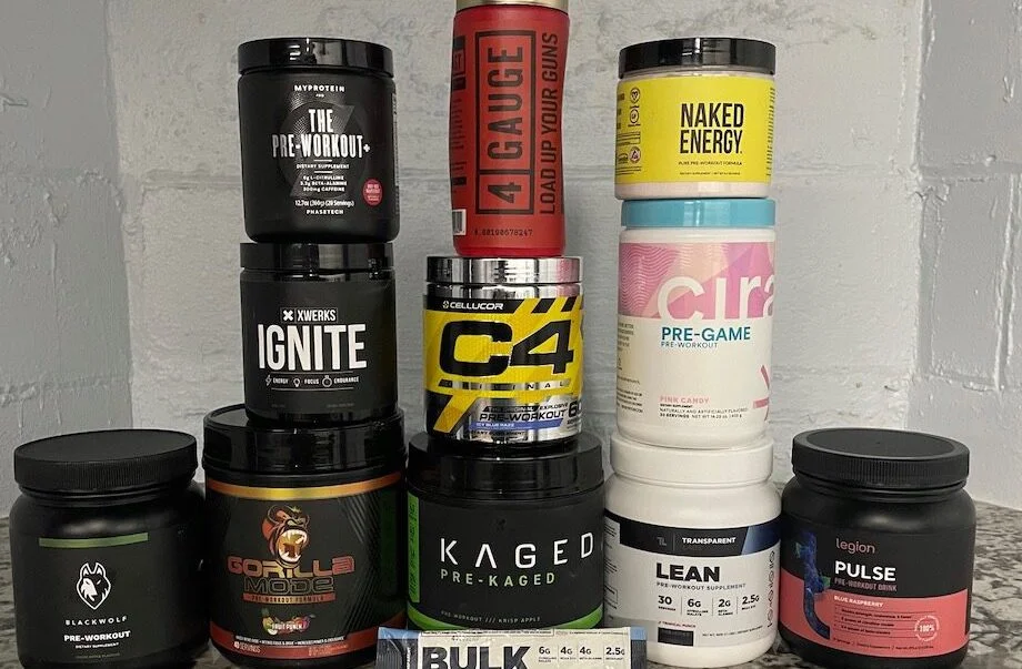 Twelve different types of the best pre-workouts we’ve tested at Garage Gym Reviews are stacked on top of each other in front of a white cinderblock wall