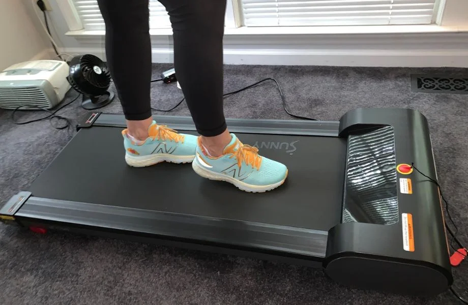 A person's feet are shown walking on the Sunny Health and Fitness Under-Desk Treadmill