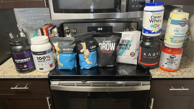 Different types of the best protein powder we’ve tested at Garage Gym Reviews in our testing facility are spread over a stove and the surrounding countertops