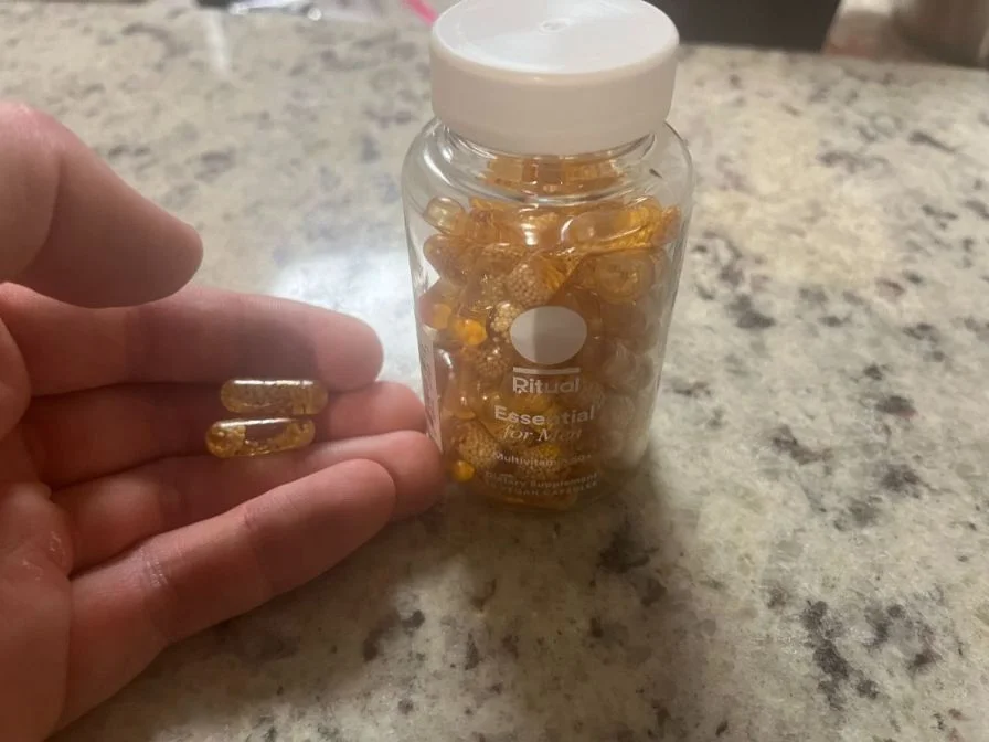 Close-up of a hand holding two clear yellowish capsules next to a clear bottle of Ritual Essential For Men Over 50.