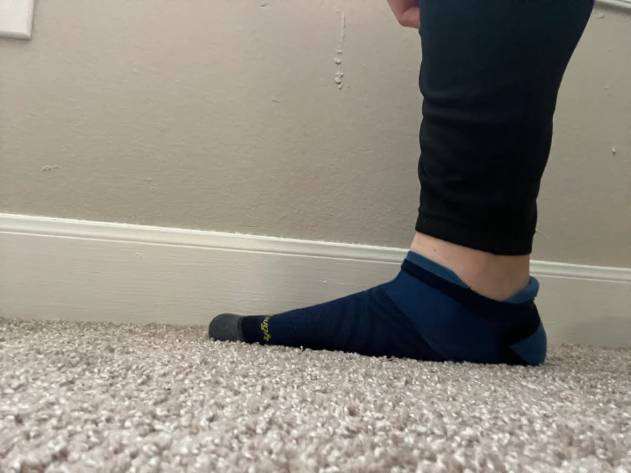 Side view of a person's foot while standing on a carpet wearing the blue and black DarnTough Men's Run No Show Tab Ultra-Lightweight Running Sock.