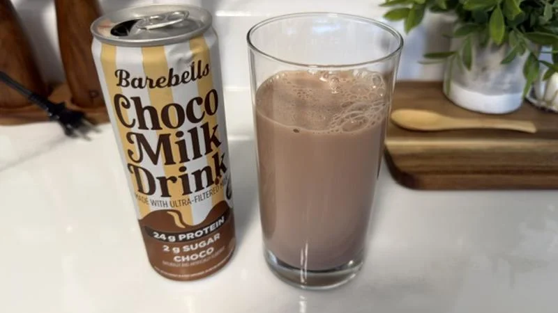 A glassful of Barebells Choco Milk sits on a white marble kitchen countertop next to the opened can. Wooden seasoning shakers can be seen out of focus in the left background, and on the right a plant and wooden spoon rest on a wooden cutting board