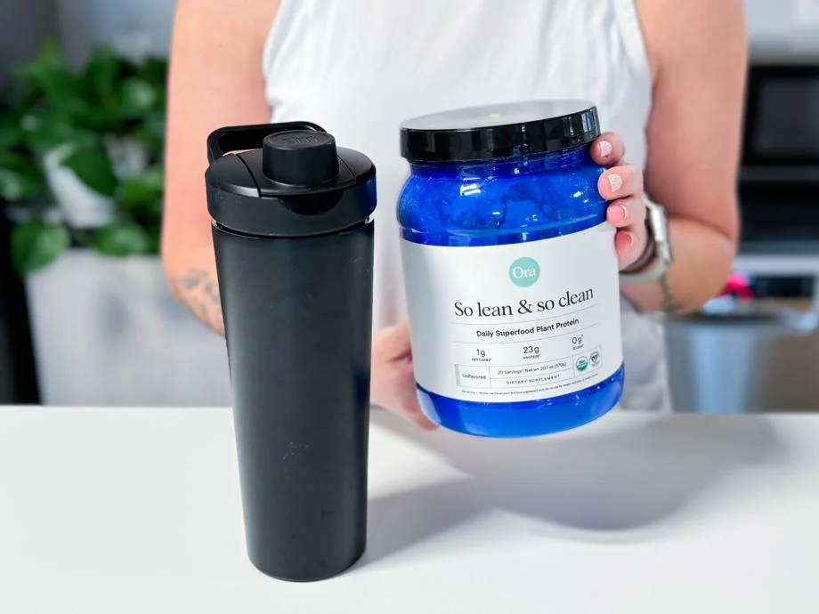 Holding a tub of Ora Organics So Lean & So Clean Plant Based Superfood Protein