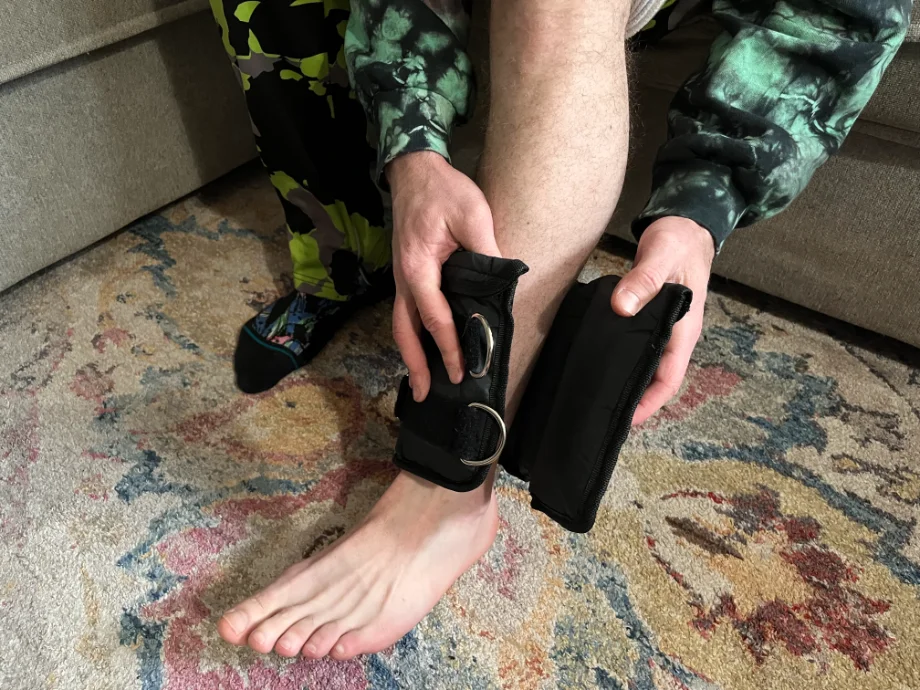Overhead shot of a man's left calf while putting on the black Synergee ankle weights with two metal loop fasteners