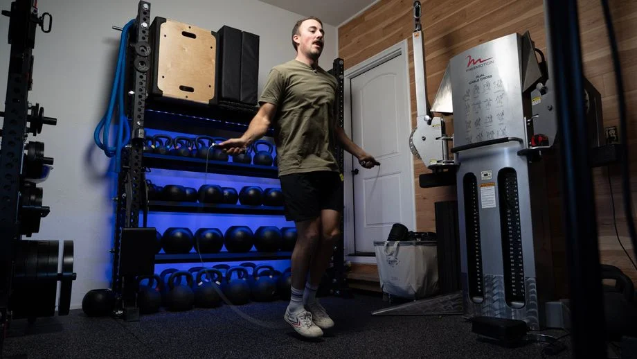 A man jumping rope in a home gym with a blue lighted kettlebell weight rack in the background.